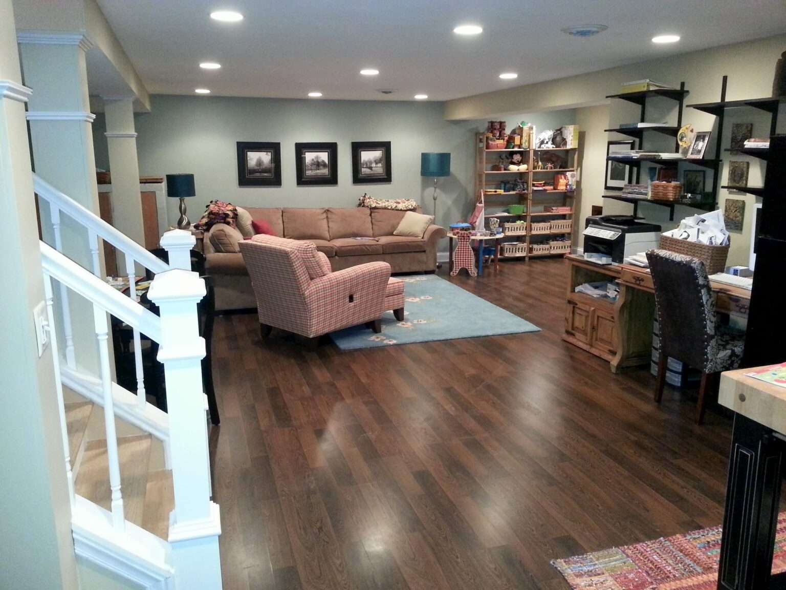 A finished basement recreation room with brown wood-look flooring, tan upholstered furniture, recessed lighting, white support columns, built-in shelving, and a staircase with white railings.