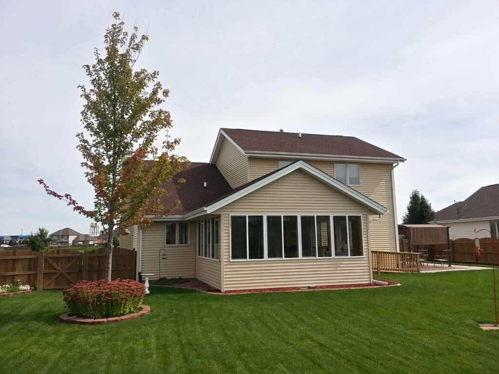 Exterior view of a two-story residential home with a new sunroom addition featuring multiple windows, vinyl siding, and a landscaped backyard with lawn and garden beds.