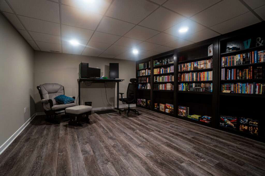 A finished basement recreation room with dark wood-look vinyl flooring, drop ceiling with recessed lighting, built-in black shelving filled with media and books, a gray gaming chair with ottoman, desk with monitor, and a comfortable upholstered armchair.