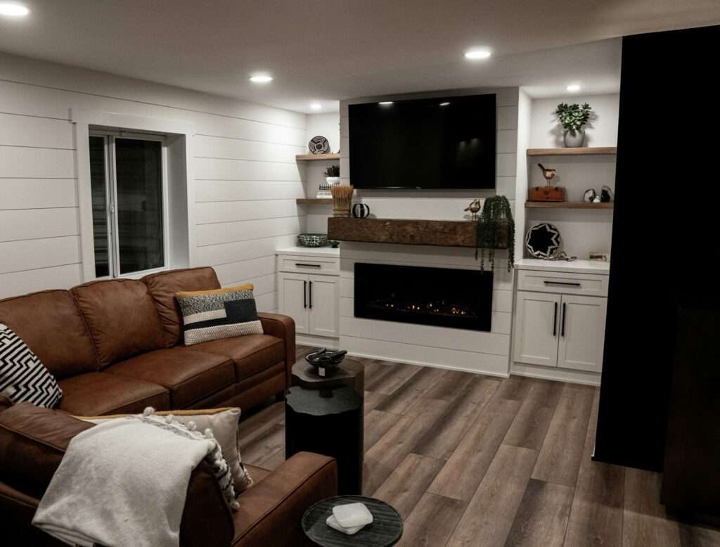 A modern living room with a brown leather sofa, built-in white cabinetry with floating shelves, a mounted flat-screen TV above an electric fireplace, and wood-look flooring.