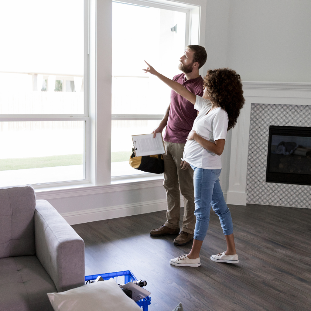 A man and woman stand in a bright, modern living room with large windows and a fireplace. The woman is pointing at something outside while the man, holding a clipboard and wearing a tool bag, looks on. A grey couch and blue basket are visible in the foreground.