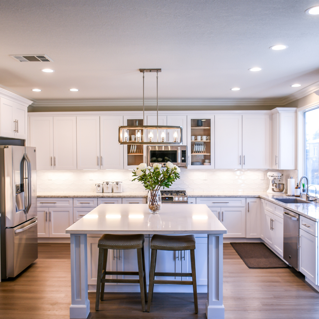 A modern kitchen with white cabinets, stainless steel appliances, and a center island featuring two barstools. The island has a vase with flowers and is lit by a stylish overhead light. The backsplash is white, and natural light fills the room from a large window—ideal inspiration for kitchen remodeling near Rockford, Illinois.