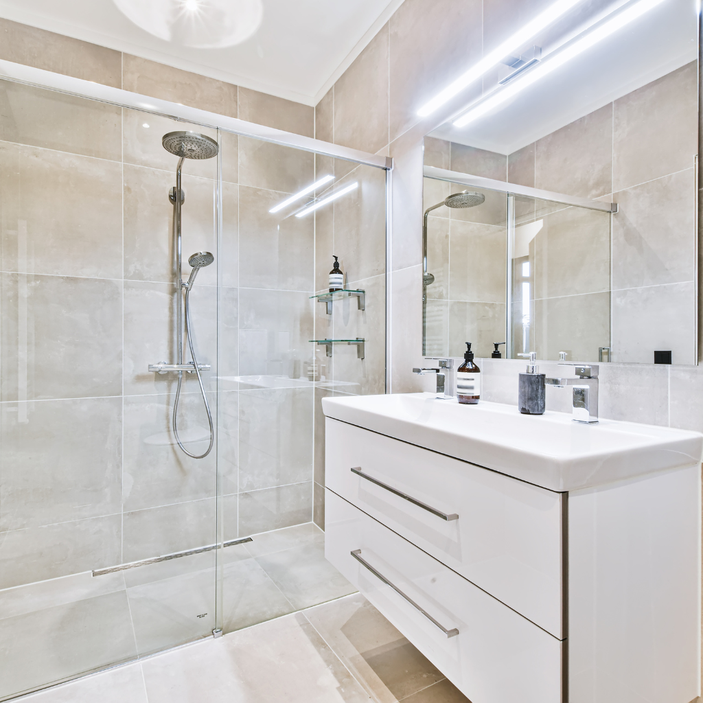 After remodeling, a modern bathroom with beige tiled walls and floors features a glass-enclosed shower, rain showerhead, and built-in shelf. Adjacent is a white vanity with a large mirror and two drawers, topped with a sink, soap dispenser, and toiletries.