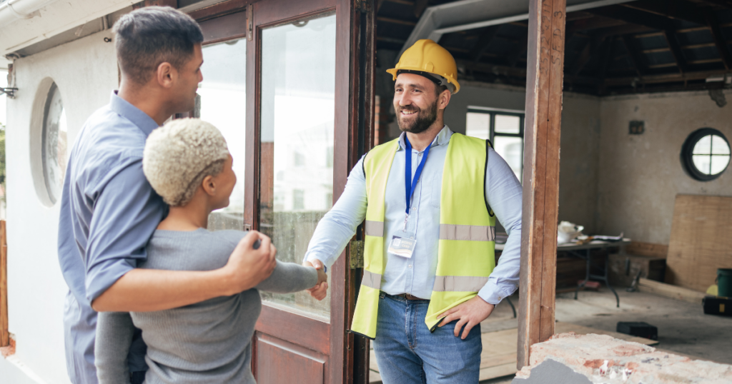 A couple stands at the entrance of a house under construction, shaking hands with a construction worker wearing a yellow hard hat and reflective vest. The man has an ID badge hanging from a lanyard. The scene is bright and welcoming—a perfect start to their home remodeling near Rockford, Illinois.