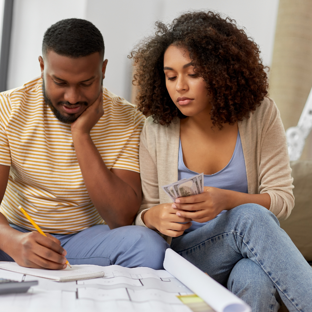 A couple sits on a sofa, focused on financial planning. The man is writing on a notebook with a pencil, while the woman, holding dollar bills, looks on thoughtfully. They seem engaged in budgeting or evaluating expenses together.