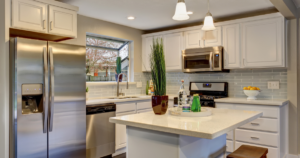 Modern kitchen with white cabinets and light blue tile backsplash. Features stainless steel appliances including a refrigerator, oven, and dishwasher. A white island counter is at the center with decorative items and pendant lights overhead.