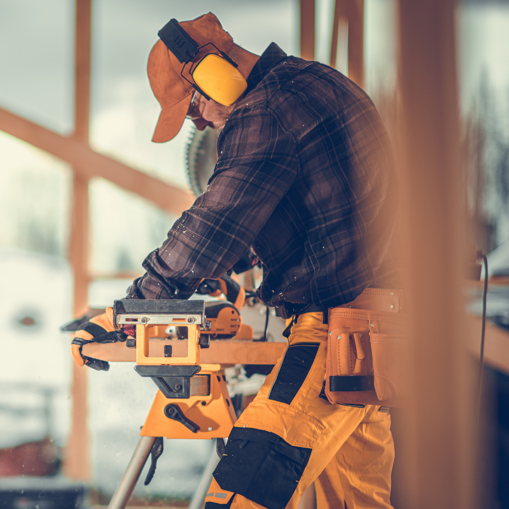 A construction worker wearing a cap and earmuffs operates a power saw at a building site. The worker is dressed in a plaid shirt and yellow pants, and wooden frames are visible in the background.