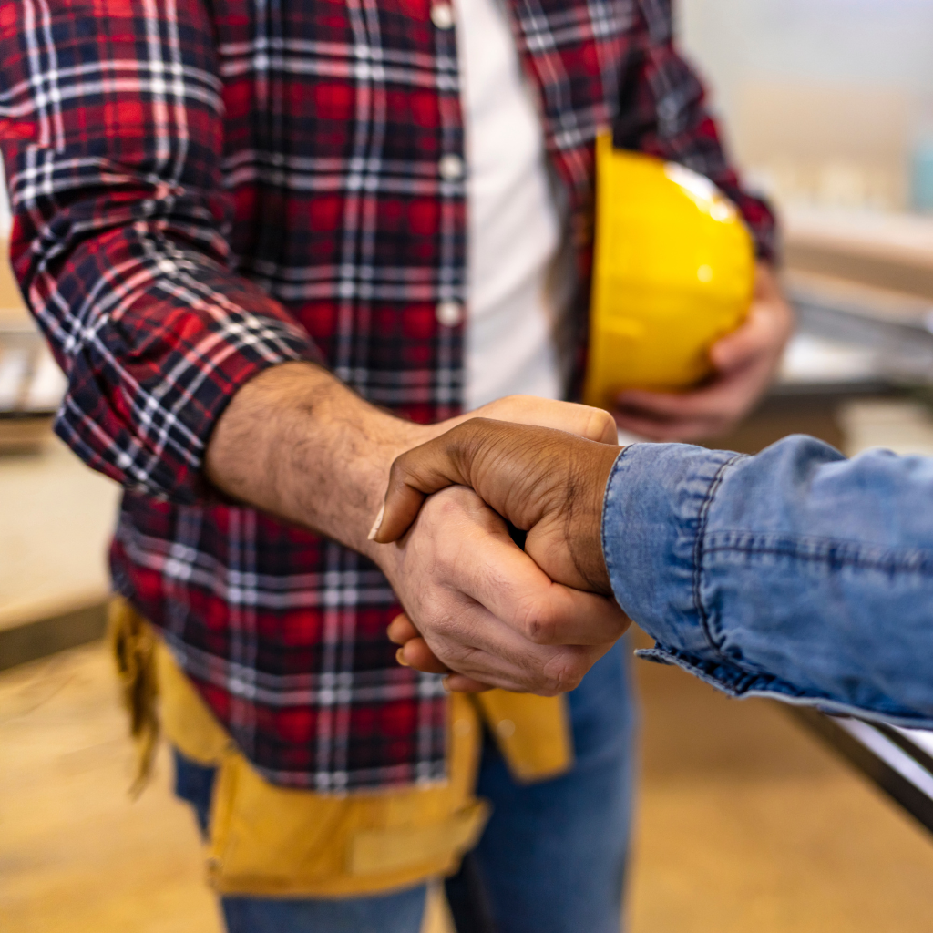 Two people shaking hands in a workshop. One wears a red plaid shirt and holds a yellow hard hat, while the other wears a denim shirt. The background features workbenches and tools, suggesting a collaborative or business agreement in a woodworking setting.