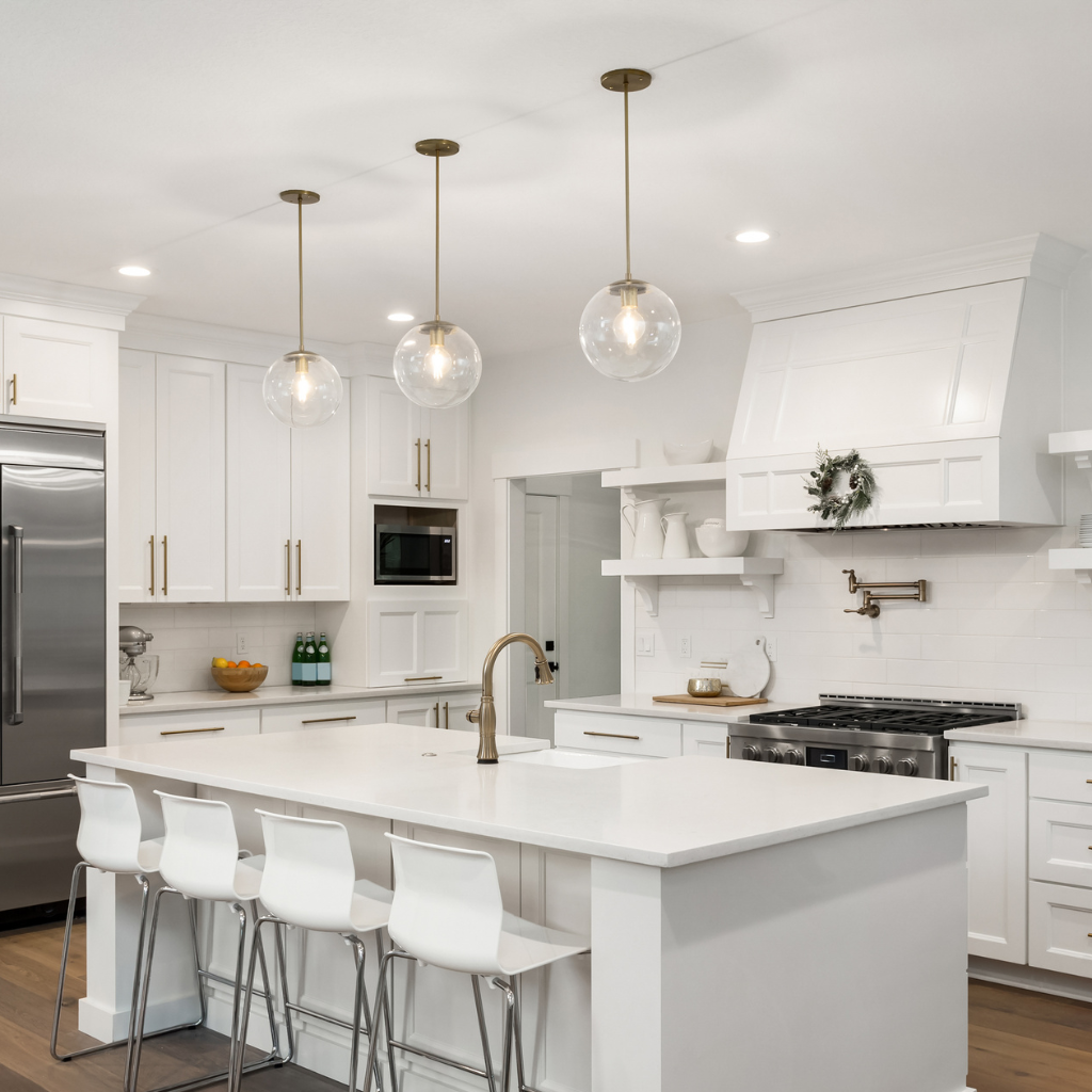 Modern kitchen with white cabinets, a large stainless steel refrigerator, and a central island with four white barstools. Pendant lights hang above the island, and a window reveals greenery outside. Shelves display decorative items.