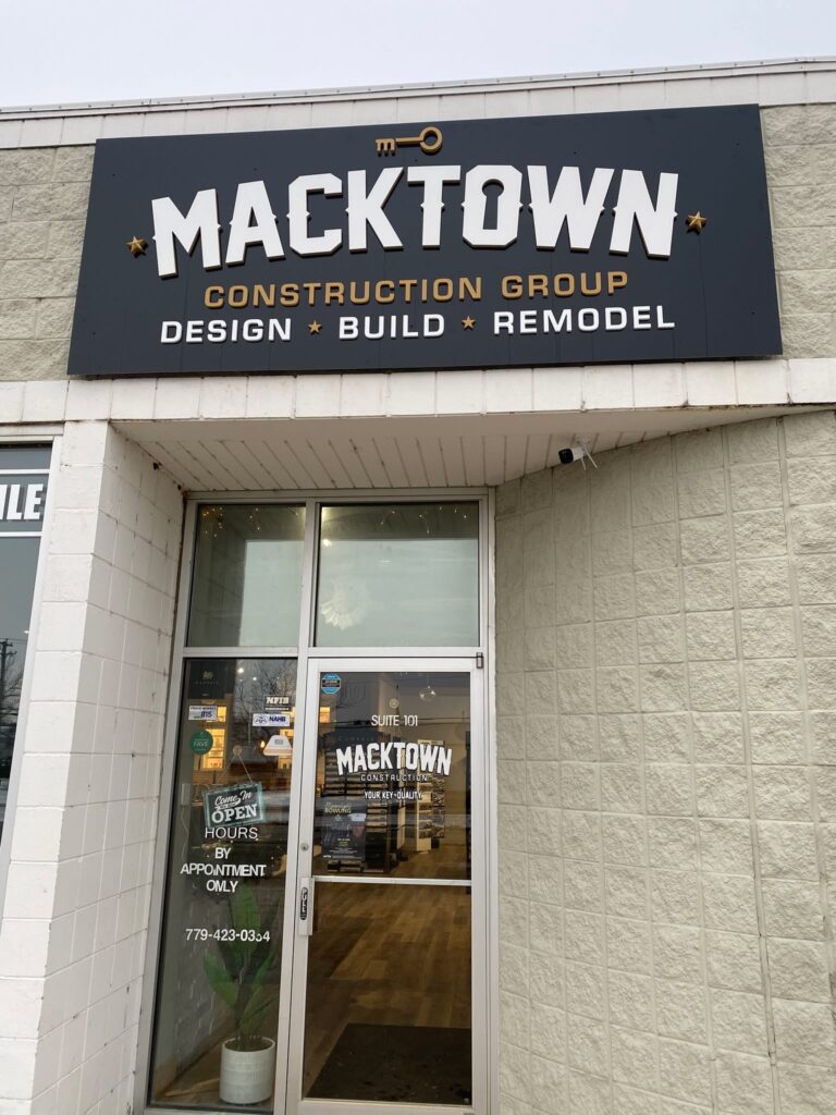 Entrance to Macktown Construction Group with a sign above reading "Design, Build, Remodel." The glass door displays the company name again, along with business hours. A potted plant is visible inside the entrance.