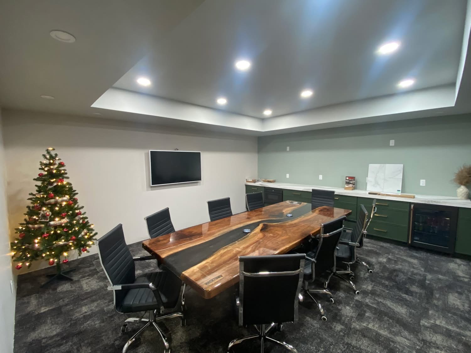 A modern conference room with a large wooden table, surrounded by black chairs. A small decorated Christmas tree stands in the corner. A flat-screen TV is mounted on the wall, and there are cabinets along the side with decor items on top.