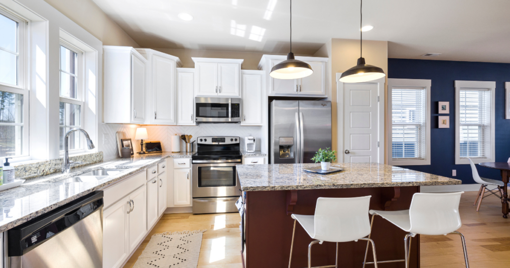 A modern kitchen features white cabinets, a stainless steel oven and refrigerator, and a granite island with bar stools. Pendant lights hang above the island. Sunlight streams through the windows, illuminating the light wood floor.