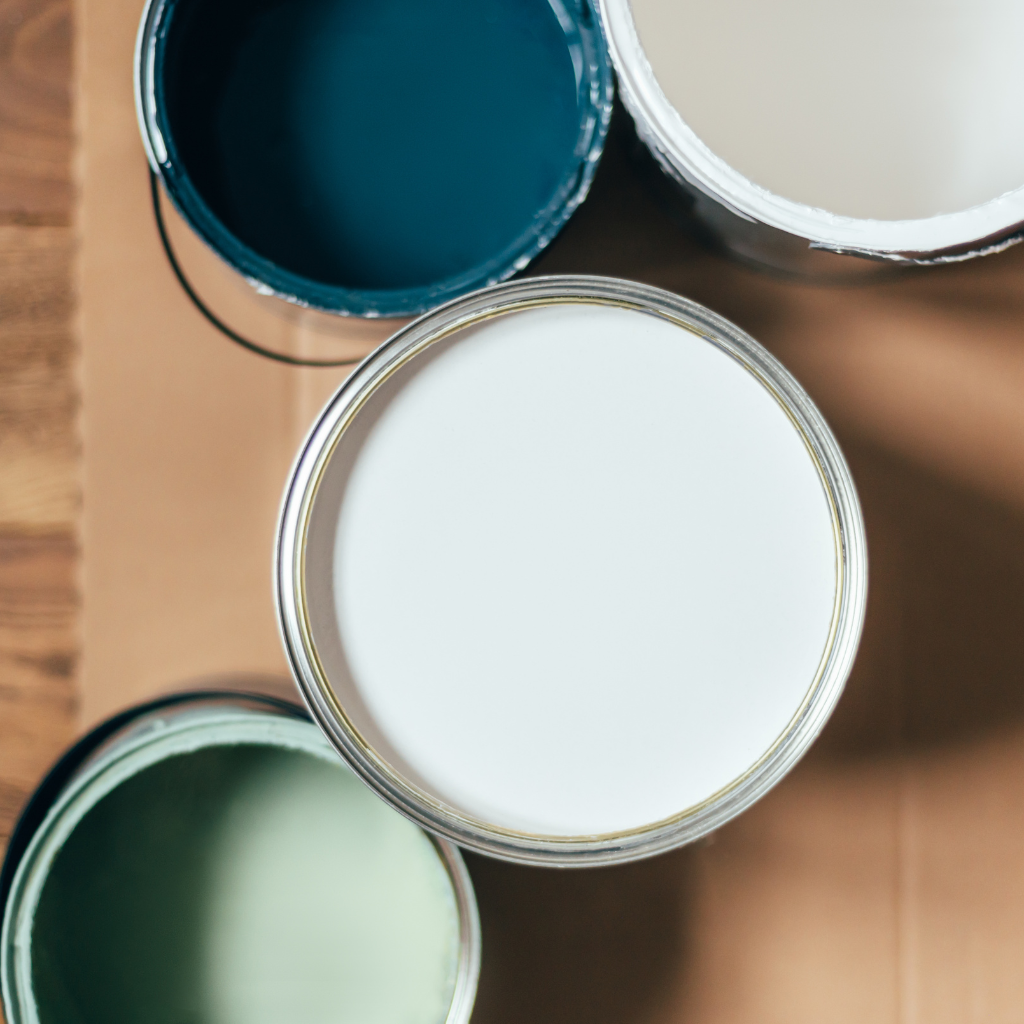 Four open paint cans are arranged on a wooden surface. The cans contain paint in shades of dark teal, white, light green, and off-white, offering a variety of color options.