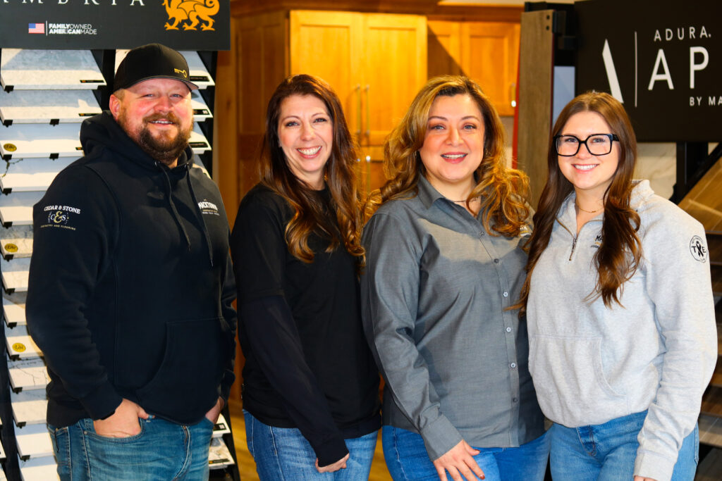 Four people standing inside a store, smiling at the camera. They are casually dressed, with two women wearing black tops and two wearing gray. A display of samples is visible on the left, and a sign with "Adura Apex" is on the right.