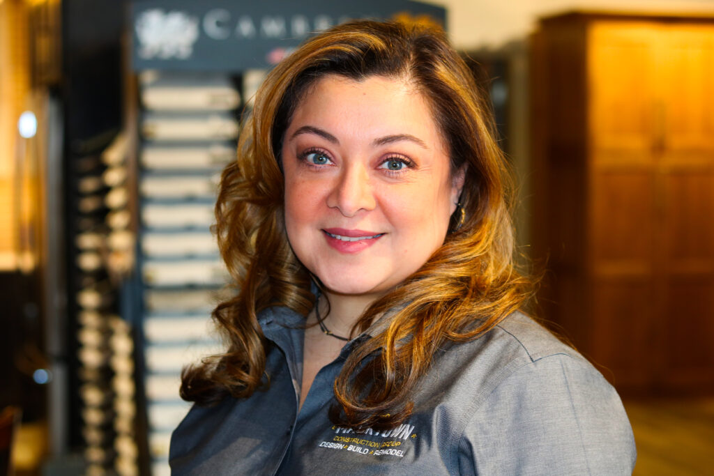 A woman with wavy, light brown hair smiles while standing indoors. She is wearing a gray shirt with text on it. The background is softly blurred, showing display boards and wooden cabinets.