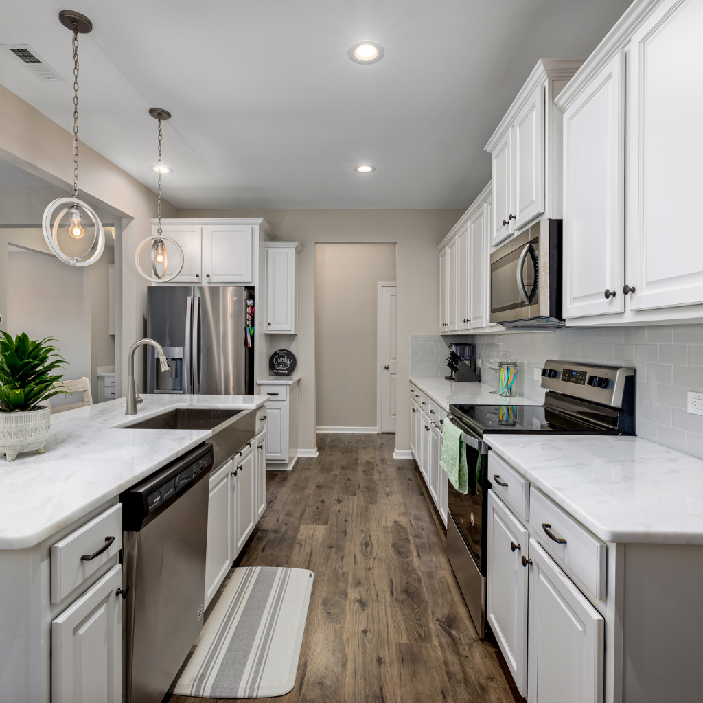 Modern kitchen with white cabinets, stainless steel appliances, and a marble countertop. There are two pendant lights, a potted plant on the island, and a striped rug on wooden flooring. The space is bright and inviting.