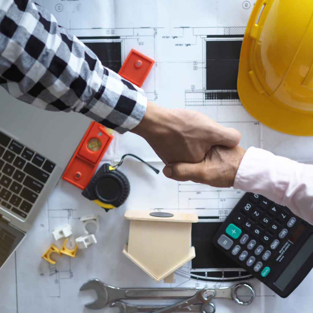 Two people shaking hands over architectural plans. There are a laptop, hard hat, calculator, measuring tape, wrench, and small model house on the table, suggesting a construction or design agreement.