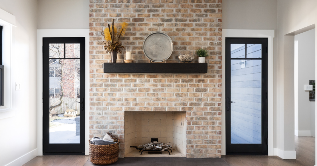 Cozy living room with a central brick fireplace, a dark wood mantel holding decorative items. Flanked by two narrow black-framed glass doors, the scene is bright and welcoming with a basket of blankets by the hearth.