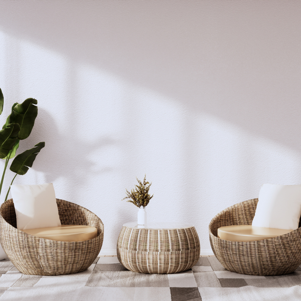 Two round wicker chairs with white cushions face a matching wicker coffee table holding a small vase with dried flowers. A large plant sits in the corner, and sunlight streams across the light-colored wall and patterned rug.