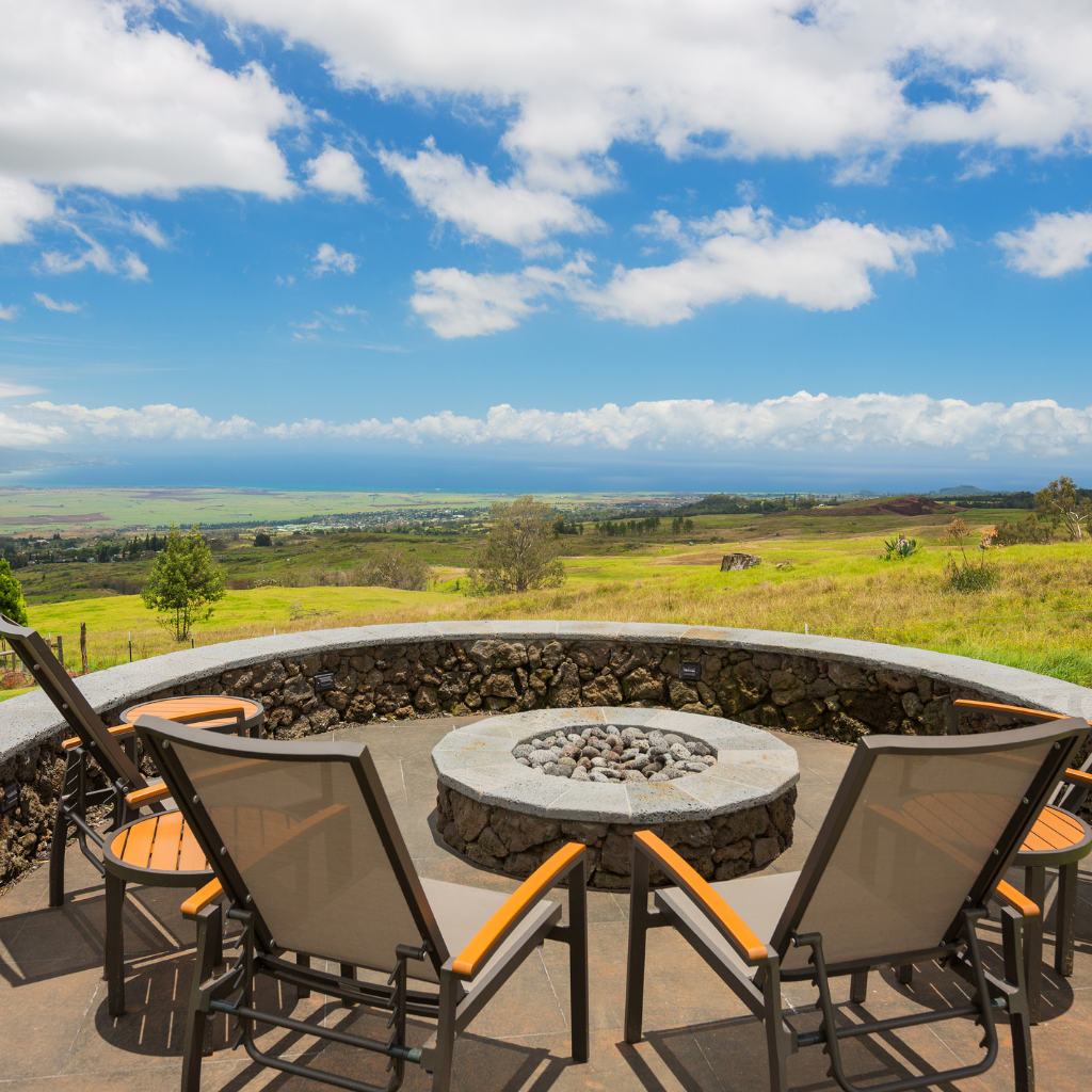 Chairs surround a stone fire pit on a patio overlooking a vast green landscape and hills under a bright blue sky with scattered clouds.
