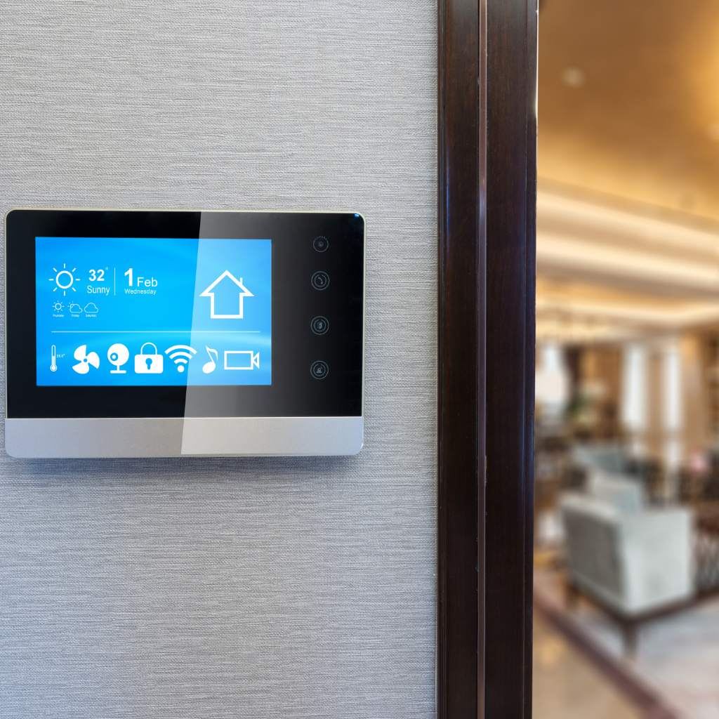 A smart home control panel mounted on a wall displays weather, date, and various home automation icons. The background shows a blurred modern living room with contemporary furniture.