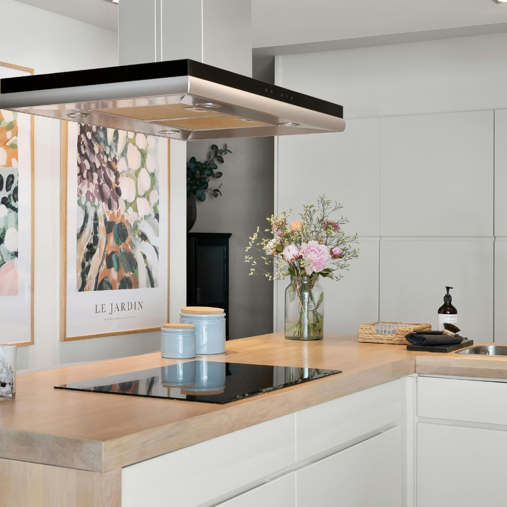 Modern kitchen with light wood countertops, white cabinets, and an electric stovetop. A vase with pink and white flowers sits next to blue canisters. Framed abstract art hangs on the wall in the background.