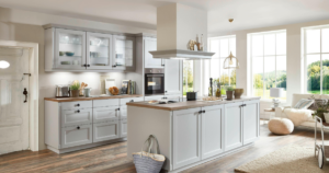 Modern, bright kitchen with light gray cabinets, wood countertops, and an island. As a trusted kitchen remodeling contractor, we designed large windows for abundant light. Appliances and plants enhance the space. Cozy living area is visible in the background.