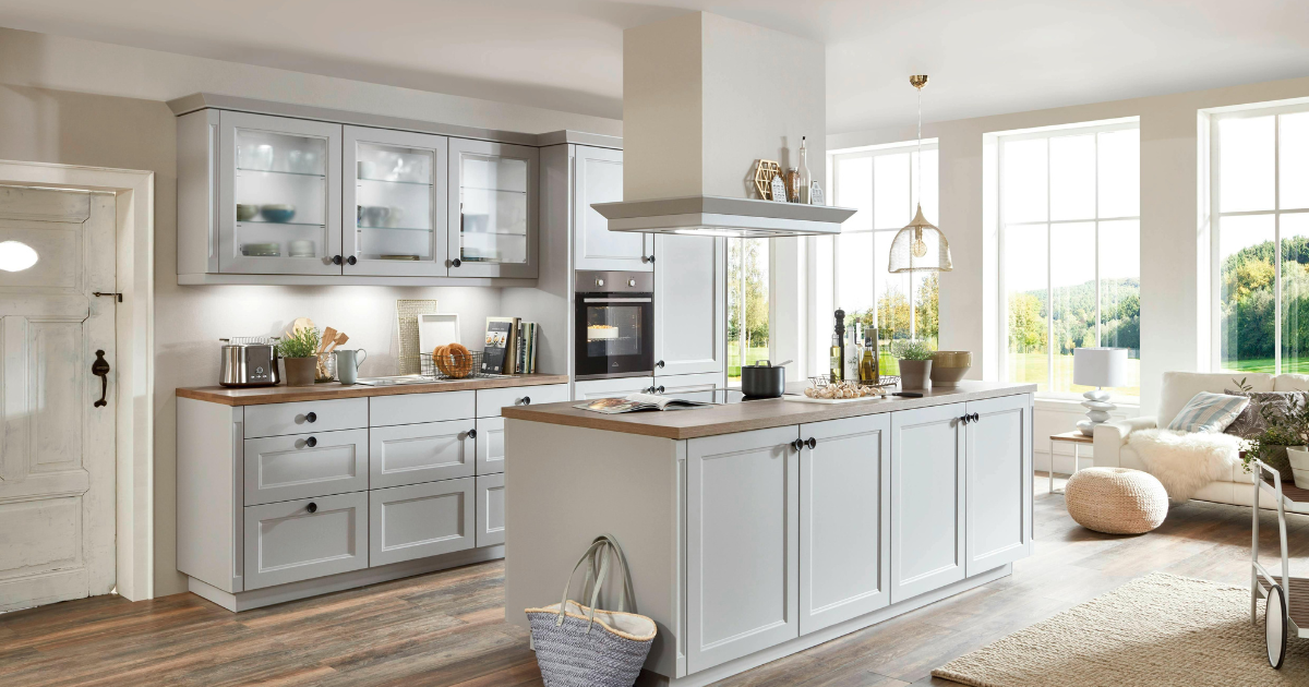 Modern, bright kitchen with light gray cabinets, wood countertops, and an island. As a trusted kitchen remodeling contractor, we designed large windows for abundant light. Appliances and plants enhance the space. Cozy living area is visible in the background.