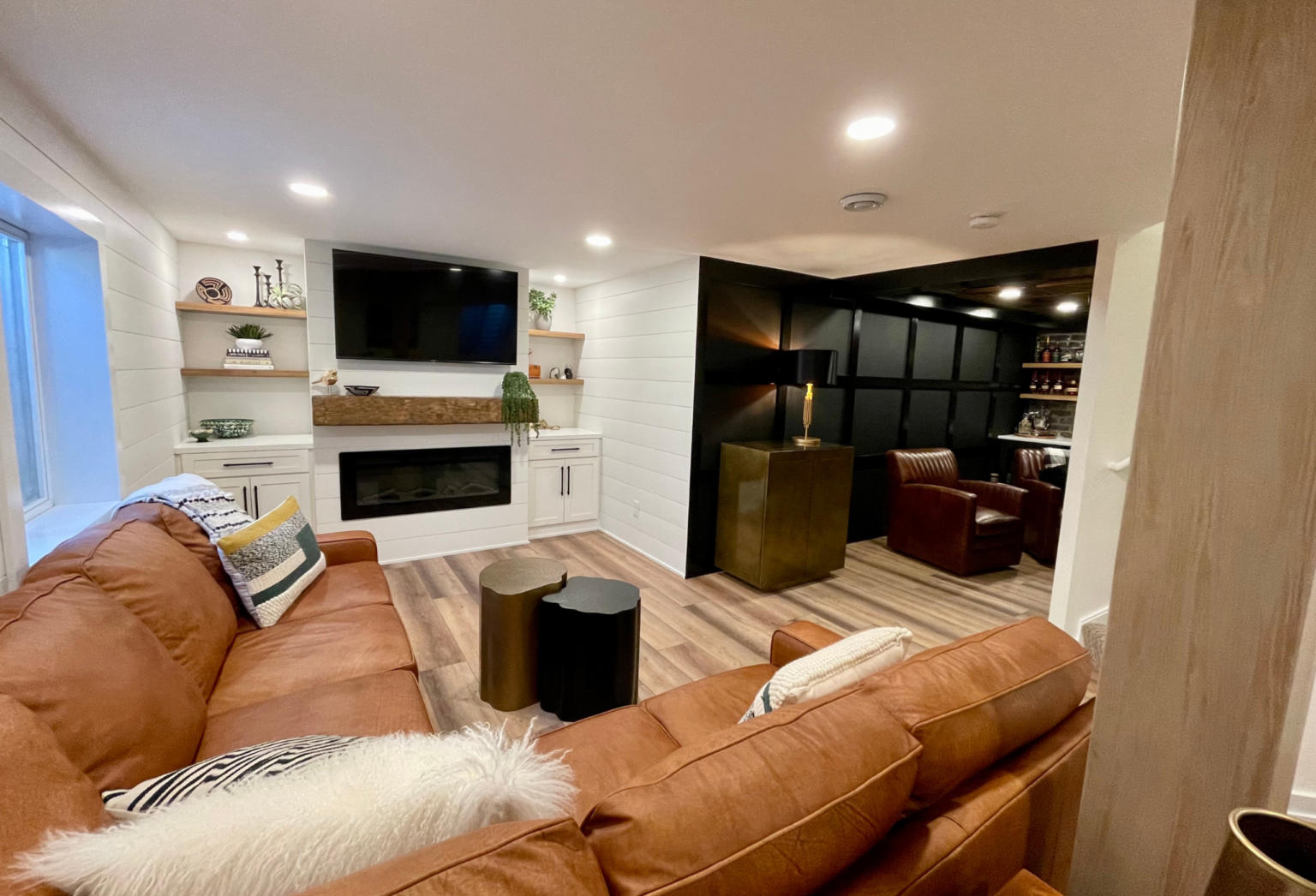 A finished basement recreation room featuring a cognac leather sectional sofa, white shiplap wall with fireplace and mounted TV, wooden flooring, recessed lighting, and a separate lounge area with black cabinetry and burgundy seating visible through an open doorway.