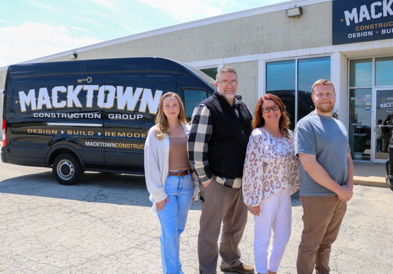 Four people stand smiling in front of a black van labeled “MACKTOWN Construction Group,” parked outside a building with the same logo and signage.